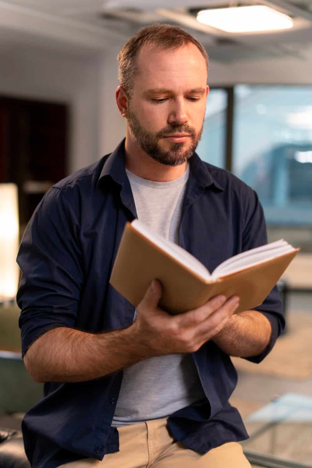 man sitting on the corner of a sofa inently reading one of the books by School of Evangelism